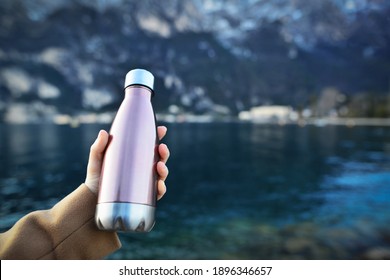 Close-up of female hand holding reusable, steel thermo shiny bottle for water, on the background of clear water of a lake with a turquoise hue. Copy space concept. 