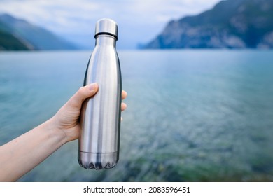 Close-up of female hand holding reusable, steel thermo shiny bottle for water, on the background of clear water of a lake with a turquoise hue. Copy space concept.