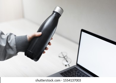 Close-up Of Female Hand Holding Steel Eco Thermo Bottle For Water Over Office Desk, Laptop With Mockup And Glasses.