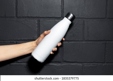 Close-up Of Female Hand, Holding Steel Reusable Thermo Water Bottle Of White Color, On Background Of Black Brick Wall.