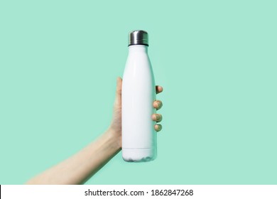Close-up of female hand, holding white reusable steel stainless eco thermo water bottle on background of cyan, aqua menthe color. Be plastic free. Zero waste. - Shutterstock ID 1862847268