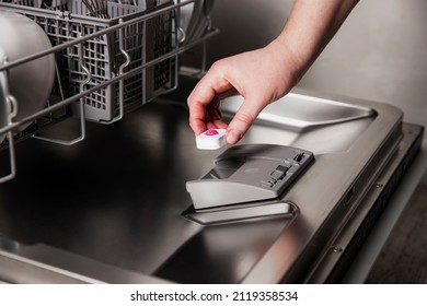 Close-up of female hand inserting dishwasher tablet into open automatic stainless built-in dishwasher machine with dirty utensils inside in modern home kitchen. Household, housekeeping domestic life