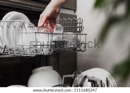 Close-up of female hand loading dished to, empty out or unloading from open automatic stainless built-in dishwasher machine with clean utensils inside in modern home kitchen. Household domestic life