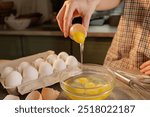 Close-up of female hands breaking an egg into a transparent bowl with eggs against the background of a wooden table. On the table are eggs in a box and a tool for making dough