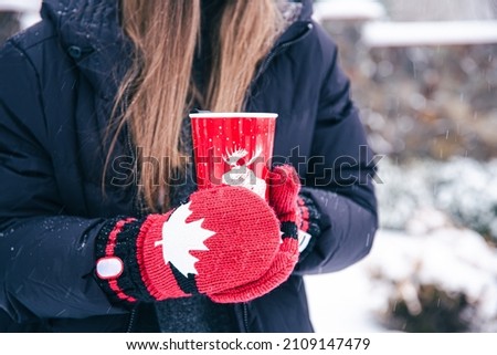 Close-up of female hands in Canada mittens hold a red thermal cup.