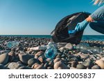 Close-up of female hands collecting plastic, rubber garbage in a black garbage bag on a pebble beach against the background of the sea with copy space. Environmental pollution on the Black Sea coast.