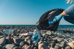 Close-up Of Female Hands Collecting Plastic, Rubber Garbage In A Black Garbage Bag On A Pebble Beach Against The Background Of The Sea With Copy Space. Environmental Pollution On The Black Sea Coast.