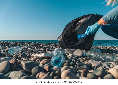 Close-up of female hands collecting plastic, rubber garbage in a black garbage bag on a pebble beach against the background of the sea with copy space. Environmental pollution on the Black Sea coast.