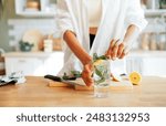 Close-up of a female hands and a glass of lemonade with mint leaves. A young woman prepares a delicious ice drink with a slice of lemon. A girl in casual clothes cuts a lemon in the kitchen at home.