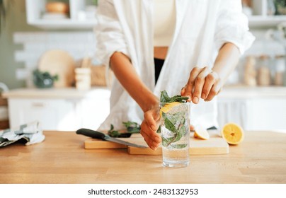 Close-up of a female hands and a glass of lemonade with mint leaves. A young woman prepares a delicious ice drink with a slice of lemon. A girl in casual clothes cuts a lemon in the kitchen at home.