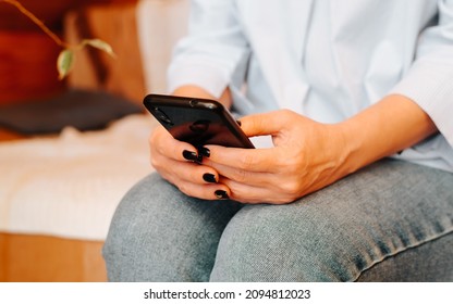 Close-up Of Female Hands Holding Black Mobile Phone, Indoors. Caucasian Woman Chatting, Typing, Browsing Internet On Smartphone While Sitting On Couch At Home. Selective Soft Focus.