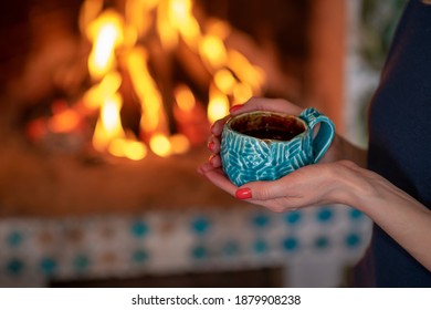 Close-up of female hands holding a cup of hot warming drink on the background of the fireplace fire. Selective focus.