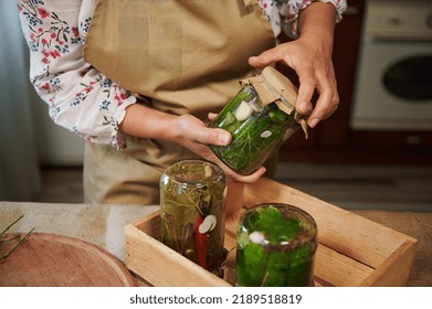 Close-up of female hands holding a glass can of pickled cucumbers and placing it upside down on a wooden crate with freshly pickled canned cucumbers and chili peppers, in the home kitchen - Shutterstock ID 2189518819