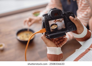 Closeup of female hands holding pro photo camera with cream soup dish on screen while doing food photography set in studio copy space - Powered by Shutterstock