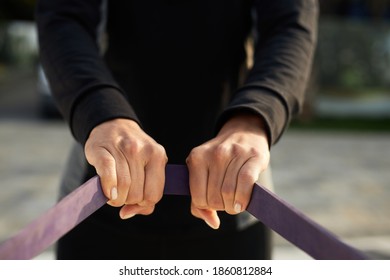 Close-up Of Female Hands Holding And Pulling Purple Fitness Elastic Band, Woman In Black Sportswear In The Background