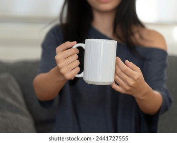 Closeup of female hands with a mug of beverage. A girl in grey t-shirt holding white mug in hand. Mug for PSD mockup. Cropped close-up image of a coffee cup. Mug for your design. Empty.