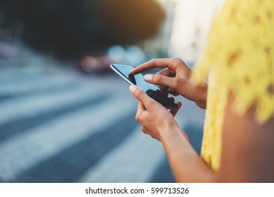 Closeup of female hands using app with city touristic map via modern smartphone device outdoors, hipster girl wearing yellow dress and typing on touch screen of cellphone while standing at crosswalk