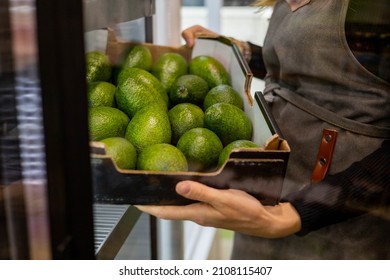 Closeup female kitchen staff hands in apron choosing fresh green avocado from cardboard box cooking at cafe kitchen. Woman arms holding ripe raw exotic fruits for healthy eating guacamole prepare - Shutterstock ID 2108115407