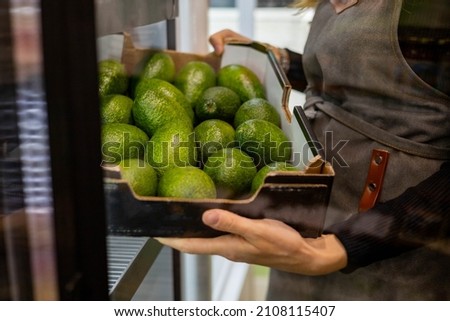 Closeup female kitchen staff hands in apron choosing fresh green avocado from cardboard box cooking at cafe kitchen. Woman arms holding ripe raw exotic fruits for healthy eating guacamole prepare