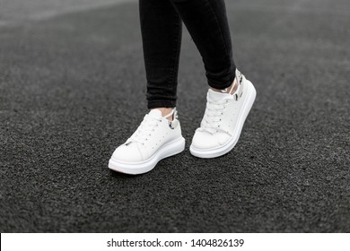 Closeup Of Female Legs In Black Jeans In Vintage Leather Sneakers On Asphalt. Fashionable Woman On A Walk. Modern Seasonal Collection Of Stylish Sneakers. Women's Fashion.