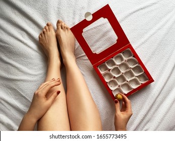 Closeup Of Female Legs On Bed With Empty Candy Box. Woman's Hands Touching The Last Chocolate Candy. Hungry Girl Eat All Candy