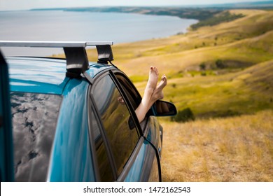 Closeup of female legs sticking out of a car window against the backdrop of the sea and hills. Summer car trip.