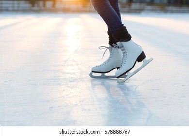 Nahaufnahme von weiblichen Beinen in weißen alten Modeschuhen auf der Eislaufbahn im Freien. Junge Frau, die auf gefrorenem See im schneebedeckten Winterpark bei Sonnenuntergang Schlittschuhlaufen