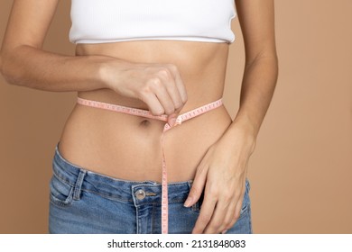 Closeup of female measuring waistline with help of tape with white bra and jeans on beige background. Weight loss. Keeping diet and exercising. Setting and achieving goals. 
