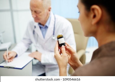 Close-up Of Female Patient Holding Bottle Of Pills And Reading Label While Senior Doctor Writing Instruction How To Take Pills In Background