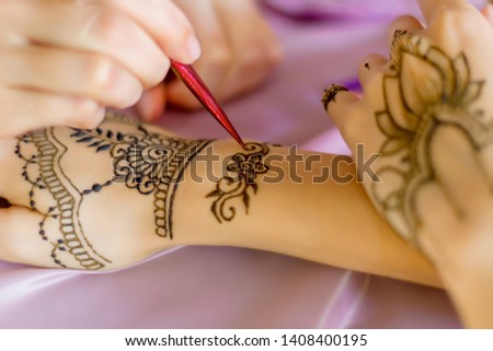 Closeup female slender wrists painted with traditional oriental mehndi ornaments. Process of painting womens hands with henna, preparing for indian wedding. Light pink fabric on background.