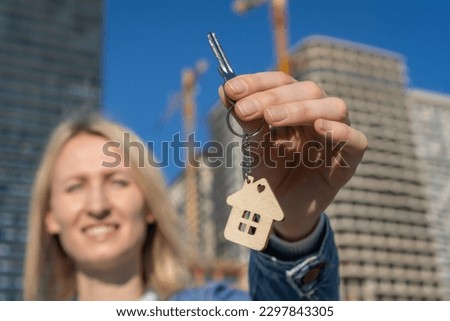 Close-up of female tenant or owner shows keys and keychain in form of house against background of newly built building - symbol of family hearth and own housing. Purchase of real estate.