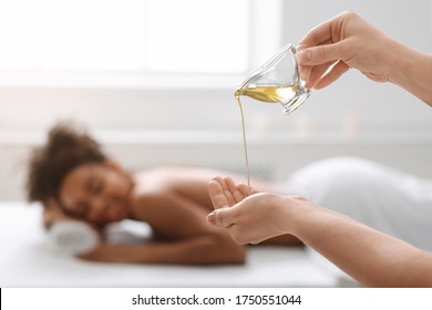 Closeup Of Female Therapist Applying Massage Oil On Hands Before Therapy, Black Lady Lying On Massage Table At Spa