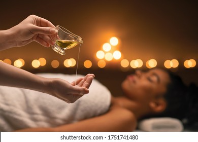 Closeup Of Female Therapist Applying Massage Oil On Hands Before Therapy, Young Black Lady Lying On Massage Table At Spa