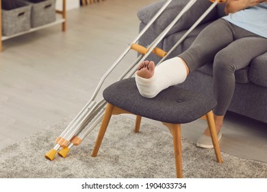 Close-up of females broken leg on stool in cast and metal crutches at home with home interior at background, selective focus. Trauma, injury, recovery, rehabilitation concept