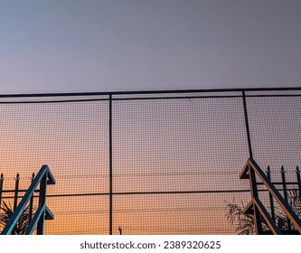 A closeup of a fence that is protecting a property under the sunset.
 The metal gate is a chain link fence with barbed wire at the very top of it. - Powered by Shutterstock