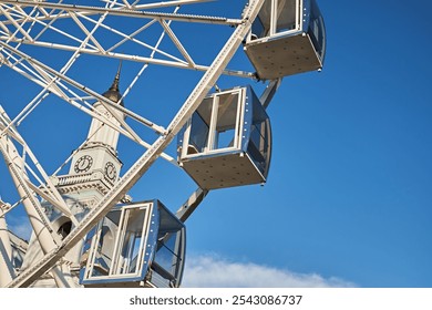 Close-up of a Ferris wheel cabin with a clock tower in the background against a clear blue sky, capturing a blend of architecture and amusement ride structure. - Powered by Shutterstock