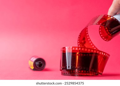 A close-up of a film reel with a strip of red film unwinding against a pink background. The film strip features perforations along the edges, showcasing a classic design - Powered by Shutterstock