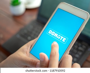 Close-up Finger Pressing Donate Icon Button On Blue Screen On White Mobile Phone With Black Laptop Computer On Wooden Table. Donation Online Concept.