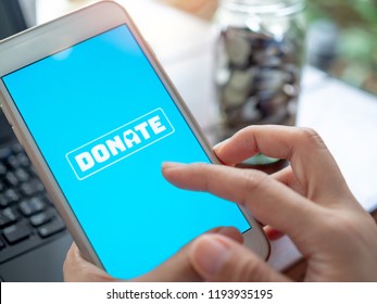Close-up Finger pressing donate icon button on blue screen on white mobile phone with coins money in glass bottle with black laptop computer. Donation online concept.
