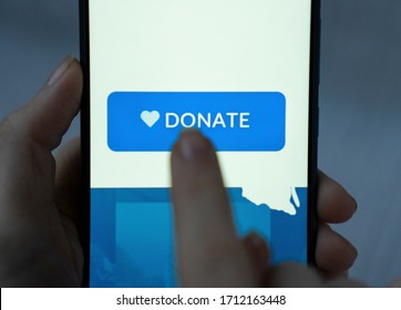 Close-up finger pressing donate icon button on blue screen on black mobile phone. Donation money online concept.