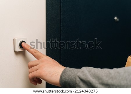 Close-up finger of unrecognizable man pushing doorbell button in entryway of apartment building. Closeup of male hand ringing door bell in block of flats. Salesman, guest or visitor behind door.