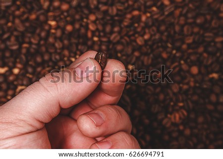 Close-up of fingers showing roasted coffee bean with blurred other coffee beans scattered behind. Shallow depth of field focused on coffee bean. Concept of individual approach to quality control.