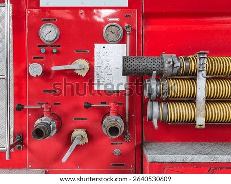 A close-up of a fire truck's control panel, featuring various gauges, valves, levers, and a map, with yellow hoses visible on the right