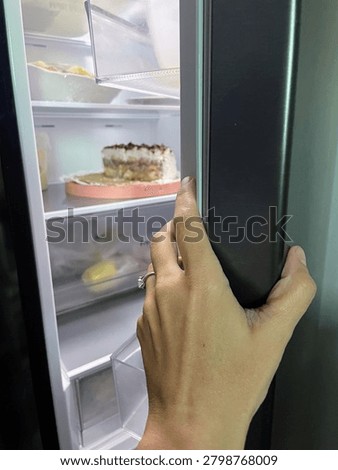 A close-up, first-person perspective shot capturing a hand with a ring opening the door of a sleek, dark-finish refrigerator. The bright interior light of the fridge illuminates the shelves, revealing a partially eaten cream cake with chocolate toppings resting on a pink platter. This image is perfect for concepts relating to late-night snacking, indulgence, kitchen appliances, food storage, or home life.