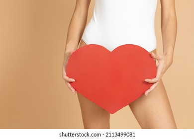 Closeup of fit skinny woman wearing white bodysuit holding big red paper heart in front of intimate area. Celebrating and congratulating loved one with Valentine's day.