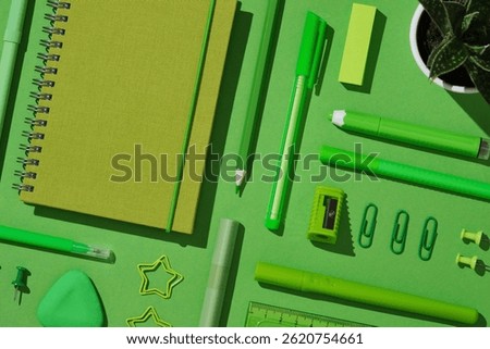 Close-up flat lay of assorted green office supplies on vibrant background, featuring spiral notebook, pens, pencil, sharpener, sticky notes, eraser, paper clips, push pins, and a small potted plant