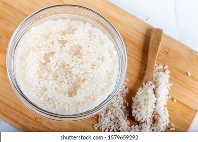 Closeup Flat Top Lay View Down Of Soaked Rice, Grain, Cloudy Liquid Water In Glass Bowl On Wooden Background