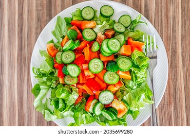 Closeup Flat Top Macro Of Fresh Raw Chopped Vegetable Salad With Romaine Lettuce Greens, Sweet Pepper, Cucumbers On White Plate, Healthy Lunch Meal By Fork On Wooden Table