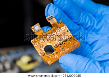 Closeup a flexible printed circuit of keypad membrane on engineer hand in blue glove. Round button switches, LED diodes or flex flat cable with connector on bent plastic board on dark blur background.