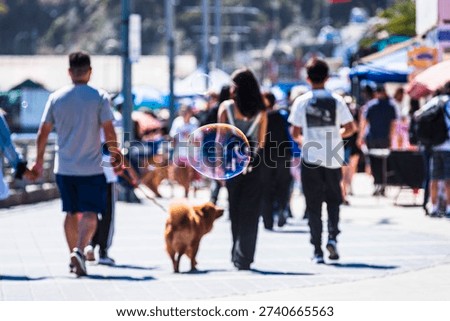 Close-up of a floating soap bubble against a bright coastal background with blurred people walking along the promenade. Sunny seaside atmosphere in San Antonio, Chile.
